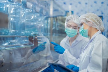 Bilim adamı, endüstriyel fabrikanın makine taşıma hattındaki su şişelerinin kalitesini kontrol ediyor. İçki üretim hattında veri kaydeden kadın işçi..