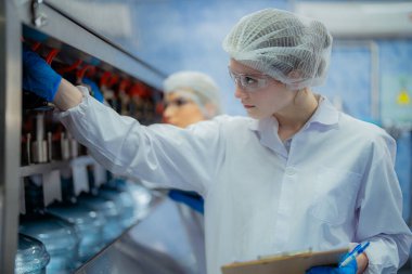 Bilim adamı, endüstriyel fabrikanın makine taşıma hattındaki su şişelerinin kalitesini kontrol ediyor. İçki üretim hattında veri kaydeden kadın işçi..