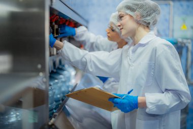 Bilim adamı, endüstriyel fabrikanın makine taşıma hattındaki su şişelerinin kalitesini kontrol ediyor. İçki üretim hattında veri kaydeden kadın işçi..