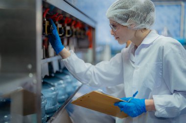 Bilim adamı, endüstriyel fabrikanın makine taşıma hattındaki su şişelerinin kalitesini kontrol ediyor. İçki üretim hattında veri kaydeden kadın işçi..