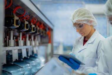 Bilim adamı, endüstriyel fabrikanın makine taşıma hattındaki su şişelerinin kalitesini kontrol ediyor. İçki üretim hattında veri kaydeden kadın işçi..