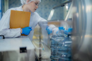 Bilim adamı, endüstriyel fabrikanın makine taşıma hattındaki su şişelerinin kalitesini kontrol ediyor. İçki üretim hattında veri kaydeden kadın işçi..