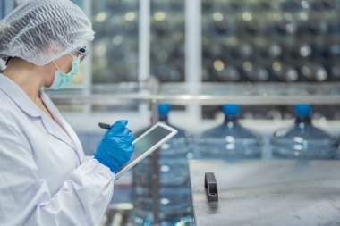 Bilim adamı, endüstriyel fabrikanın makine taşıma hattındaki su şişelerinin kalitesini kontrol ediyor. İçki üretim hattında veri kaydeden kadın işçi..