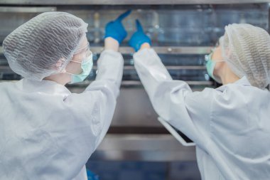 Bilim adamı, endüstriyel fabrikanın makine taşıma hattındaki su şişelerinin kalitesini kontrol ediyor. İçki üretim hattında veri kaydeden kadın işçi..