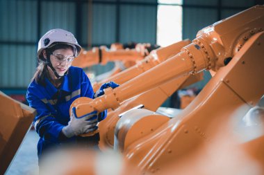 Fabrika mühendisi kadın akıllı tabletle makineyi inceliyor. İşçi robot kolunda çalışıyor. Endüstriyel bir fabrikada uzaktan kumandalı kaynak makinesi. Yapay zeka kavramı.