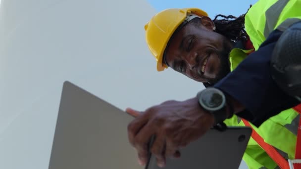 アフリカ人労働者は 風力タービンの前に青いワーキングスーツのドレスと安全ヘルメットに自信を持って座っている 再生可能エネルギーのスマート産業労働者のコンセプト — ストック動画