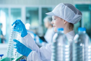 Bilim adamı, endüstriyel fabrikanın makine taşıma hattındaki su şişelerinin kalitesini kontrol ediyor. İçki üretim hattında veri kaydeden kadın işçi..