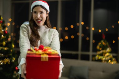 Yeni yılı kutlayan kırmızı hediye kutularını tutan güzel, gülümseyen genç Asyalı kadın. Noel hediyeleri. Beyaz örgü süveter ve şapka giyen ve Noel ağaçlarıyla poz veren bir kadın. Bir kış modası trendi.