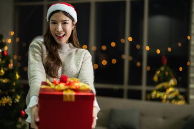 Yeni yılı kutlayan kırmızı hediye kutularını tutan güzel, gülümseyen genç Asyalı kadın. Noel hediyeleri. Beyaz örgü süveter ve şapka giyen ve Noel ağaçlarıyla poz veren bir kadın. Bir kış modası trendi.