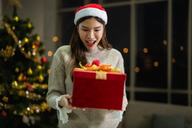 Yeni yılı kutlayan kırmızı hediye kutularını tutan güzel, gülümseyen genç Asyalı kadın. Noel hediyeleri. Beyaz örgü süveter ve şapka giyen ve Noel ağaçlarıyla poz veren bir kadın. Bir kış modası trendi.