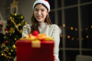 Yeni yılı kutlayan kırmızı hediye kutularını tutan güzel, gülümseyen genç Asyalı kadın. Noel hediyeleri. Beyaz örgü süveter ve şapka giyen ve Noel ağaçlarıyla poz veren bir kadın. Bir kış modası trendi.