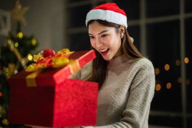 Yeni yılı kutlayan kırmızı hediye kutularını tutan güzel, gülümseyen genç Asyalı kadın. Noel hediyeleri. Beyaz örgü süveter ve şapka giyen ve Noel ağaçlarıyla poz veren bir kadın. Bir kış modası trendi.