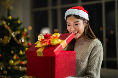 Yeni yılı kutlayan kırmızı hediye kutularını tutan güzel, gülümseyen genç Asyalı kadın. Noel hediyeleri. Beyaz örgü süveter ve şapka giyen ve Noel ağaçlarıyla poz veren bir kadın. Bir kış modası trendi.