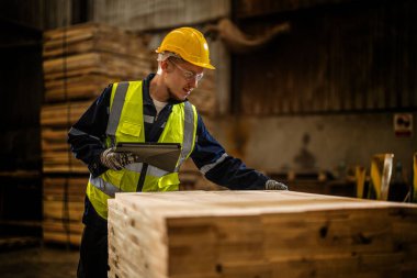 Takım çalışanı marangoz, güvenlik üniforması ve şapka takıyor atölyedeki ahşap ürünlerin kalitesini kontrol ediyor. Erkek ve kadın işçiler karanlık depo endüstrisinde ahşap..