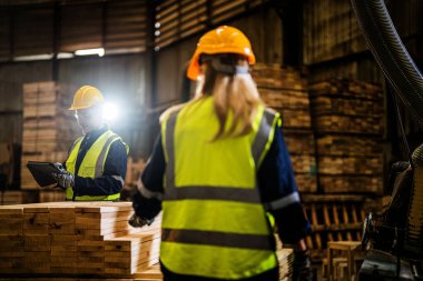 Takım çalışanı marangoz, güvenlik üniforması ve şapka takıyor atölyedeki ahşap ürünlerin kalitesini kontrol ediyor. Erkek ve kadın işçiler karanlık depo endüstrisinde ahşap..