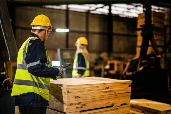 Team worker carpenter wearing safety uniform and hard hat working and checking the quality of wooden products at workshop manufacturing. man and woman workers wood in dark warehouse industry.