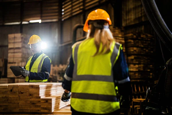 Team worker carpenter wearing safety uniform and hard hat working and checking the quality of wooden products at workshop manufacturing. man and woman workers wood in dark warehouse industry.