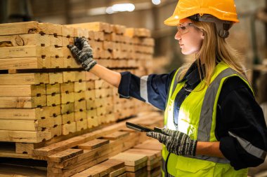 Marangoz kadın güvenlik üniforması ve şapka takıyor atölyedeki ahşap ürünlerin kalitesini kontrol ediyor. Erkek ve kadın işçiler karanlık depo endüstrisinde ahşap..