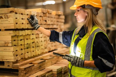 Marangoz kadın güvenlik üniforması ve şapka takıyor atölyedeki ahşap ürünlerin kalitesini kontrol ediyor. Erkek ve kadın işçiler karanlık depo endüstrisinde ahşap..