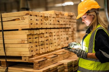 Marangoz kadın güvenlik üniforması ve şapka takıyor atölyedeki ahşap ürünlerin kalitesini kontrol ediyor. Erkek ve kadın işçiler karanlık depo endüstrisinde ahşap..