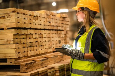 Marangoz kadın güvenlik üniforması ve şapka takıyor atölyedeki ahşap ürünlerin kalitesini kontrol ediyor. Erkek ve kadın işçiler karanlık depo endüstrisinde ahşap..