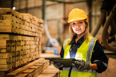 Marangoz kadın güvenlik üniforması ve şapka takıyor atölyedeki ahşap ürünlerin kalitesini kontrol ediyor. Erkek ve kadın işçiler karanlık depo endüstrisinde ahşap..