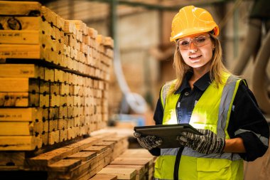 Marangoz kadın güvenlik üniforması ve şapka takıyor atölyedeki ahşap ürünlerin kalitesini kontrol ediyor. Erkek ve kadın işçiler karanlık depo endüstrisinde ahşap..