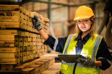 Marangoz kadın güvenlik üniforması ve şapka takıyor atölyedeki ahşap ürünlerin kalitesini kontrol ediyor. Erkek ve kadın işçiler karanlık depo endüstrisinde ahşap..