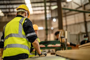 Karanlık depo endüstrisinde kereste temizleyen bir adam. Takım çalışanı marangoz, güvenlik üniforması ve şapka takıyor atölyede ahşap ürünlerin kalitesini kontrol ediyor ve çalışıyor..