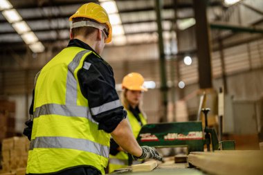 Karanlık depo endüstrisinde kereste temizleyen bir adam. Takım çalışanı marangoz, güvenlik üniforması ve şapka takıyor atölyede ahşap ürünlerin kalitesini kontrol ediyor ve çalışıyor..