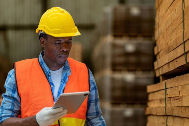 Afrikalı marangoz, güvenlik üniforması ve sıkı bir şapka giyip atölyede ahşap ürünlerin kalitesini kontrol ediyor. Erkek ve kadın işçiler karanlık depo endüstrisinde ahşap..