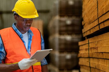 Afrikalı marangoz, güvenlik üniforması ve sıkı bir şapka giyip atölyede ahşap ürünlerin kalitesini kontrol ediyor. Erkek ve kadın işçiler karanlık depo endüstrisinde ahşap..