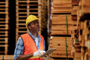 Afrikalı marangoz, güvenlik üniforması ve sıkı bir şapka giyip atölyede ahşap ürünlerin kalitesini kontrol ediyor. Erkek ve kadın işçiler karanlık depo endüstrisinde ahşap..