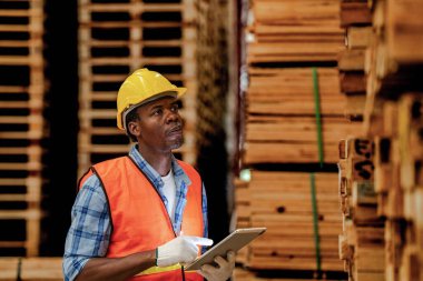 Afrikalı marangoz, güvenlik üniforması ve sıkı bir şapka giyip atölyede ahşap ürünlerin kalitesini kontrol ediyor. Erkek ve kadın işçiler karanlık depo endüstrisinde ahşap..