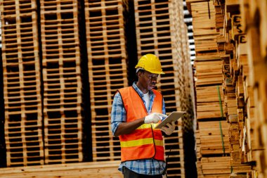 Afrikalı marangoz, güvenlik üniforması ve sıkı bir şapka giyip atölyede ahşap ürünlerin kalitesini kontrol ediyor. Erkek ve kadın işçiler karanlık depo endüstrisinde ahşap..