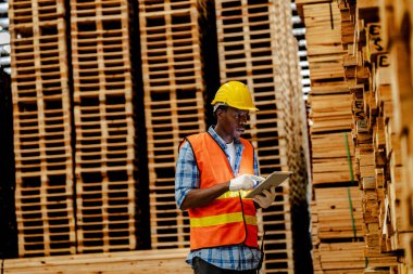 Afrikalı marangoz, güvenlik üniforması ve sıkı bir şapka giyip atölyede ahşap ürünlerin kalitesini kontrol ediyor. Erkek ve kadın işçiler karanlık depo endüstrisinde ahşap..