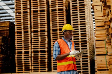 Afrikalı marangoz, güvenlik üniforması ve sıkı bir şapka giyip atölyede ahşap ürünlerin kalitesini kontrol ediyor. Erkek ve kadın işçiler karanlık depo endüstrisinde ahşap..