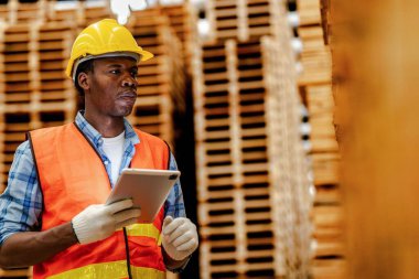 Afrikalı marangoz, güvenlik üniforması ve sıkı bir şapka giyip atölyede ahşap ürünlerin kalitesini kontrol ediyor. Erkek ve kadın işçiler karanlık depo endüstrisinde ahşap..