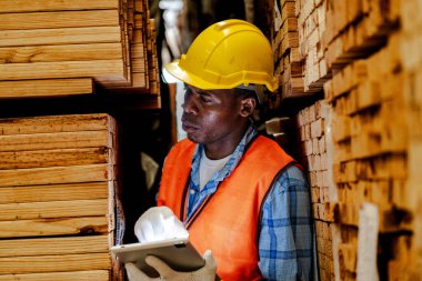 Afrikalı marangoz, güvenlik üniforması ve sıkı bir şapka giyip atölyede ahşap ürünlerin kalitesini kontrol ediyor. Erkek ve kadın işçiler karanlık depo endüstrisinde ahşap..