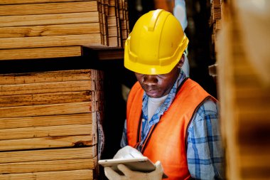 Afrikalı marangoz, güvenlik üniforması ve sıkı bir şapka giyip atölyede ahşap ürünlerin kalitesini kontrol ediyor. Erkek ve kadın işçiler karanlık depo endüstrisinde ahşap..
