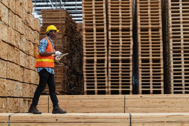 Afrikalı işçiler, kereste deposunda çalışan süit elbise ve eldiven ile yürüyen ve denetleyen mühendisler. Akıllı endüstri çalışanı kavramı. Ahşap fabrikaları ağaç damak üretir..