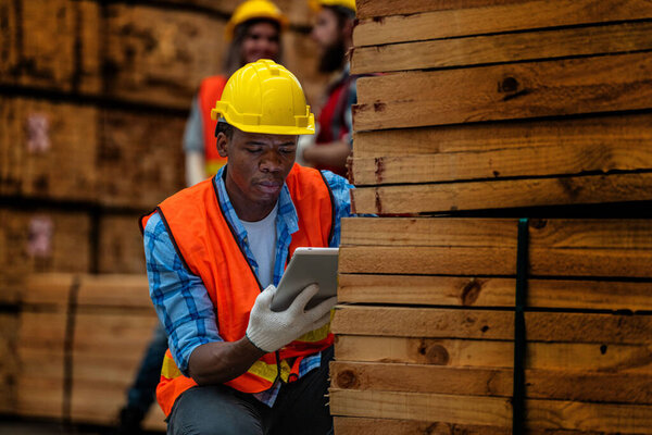 African worker carpenter wearing safety uniform and hard hat working and checking the quality of wooden products at workshop manufacturing. man and woman workers wood in dark warehouse industry.