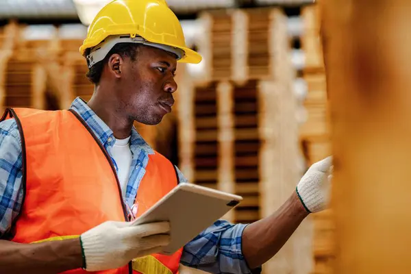 African worker carpenter wearing safety uniform and hard hat working and checking the quality of wooden products at workshop manufacturing. man and woman workers wood in dark warehouse industry.
