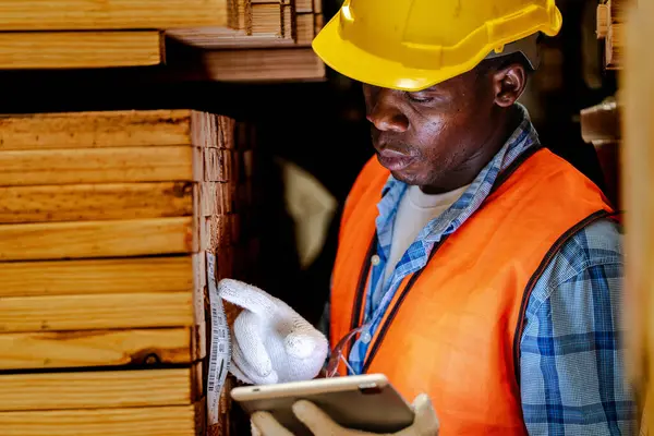 African worker carpenter wearing safety uniform and hard hat working and checking the quality of wooden products at workshop manufacturing. man and woman workers wood in dark warehouse industry.