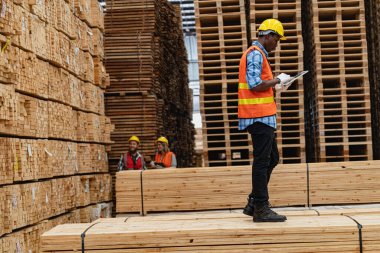 Afrikalı işçiler, kereste deposunda çalışan süit elbise ve eldiven ile yürüyen ve denetleyen mühendisler. Akıllı endüstri çalışanı kavramı. Ahşap fabrikaları ağaç damak üretir..