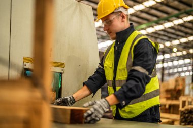 Karanlık depo endüstrisinde kereste temizleyen bir adam. Takım çalışanı marangoz, güvenlik üniforması ve şapka takıyor atölyede ahşap ürünlerin kalitesini kontrol ediyor ve çalışıyor..