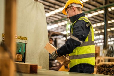 Karanlık depo endüstrisinde kereste temizleyen bir adam. Takım çalışanı marangoz, güvenlik üniforması ve şapka takıyor atölyede ahşap ürünlerin kalitesini kontrol ediyor ve çalışıyor..