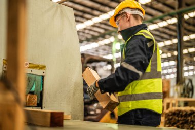 Karanlık depo endüstrisinde kereste temizleyen bir adam. Takım çalışanı marangoz, güvenlik üniforması ve şapka takıyor atölyede ahşap ürünlerin kalitesini kontrol ediyor ve çalışıyor..