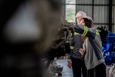 Fabrika mühendisi kadın akıllı tabletle makineyi inceliyor. İşçi robot kolunda çalışıyor. Endüstriyel bir fabrikada uzaktan kumandalı kaynak makinesi. Yapay zeka kavramı.