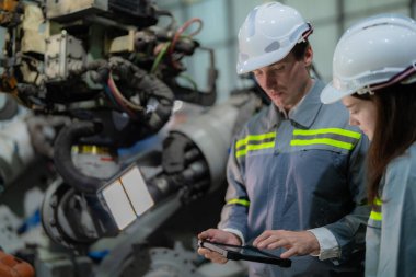 Fabrika mühendisi kadın akıllı tabletle makineyi inceliyor. İşçi robot kolunda çalışıyor. Endüstriyel bir fabrikada uzaktan kumandalı kaynak makinesi. Yapay zeka kavramı.